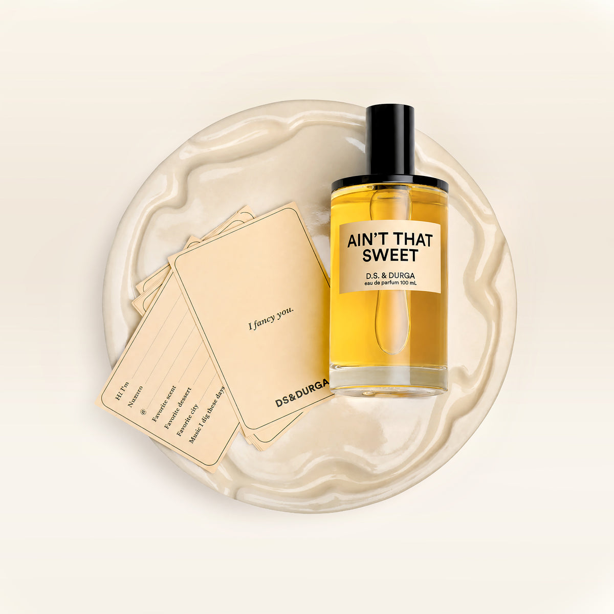 A 100 mL bottle of Ain't That Sweet perfume and flirt cards on a ceramic cake plate